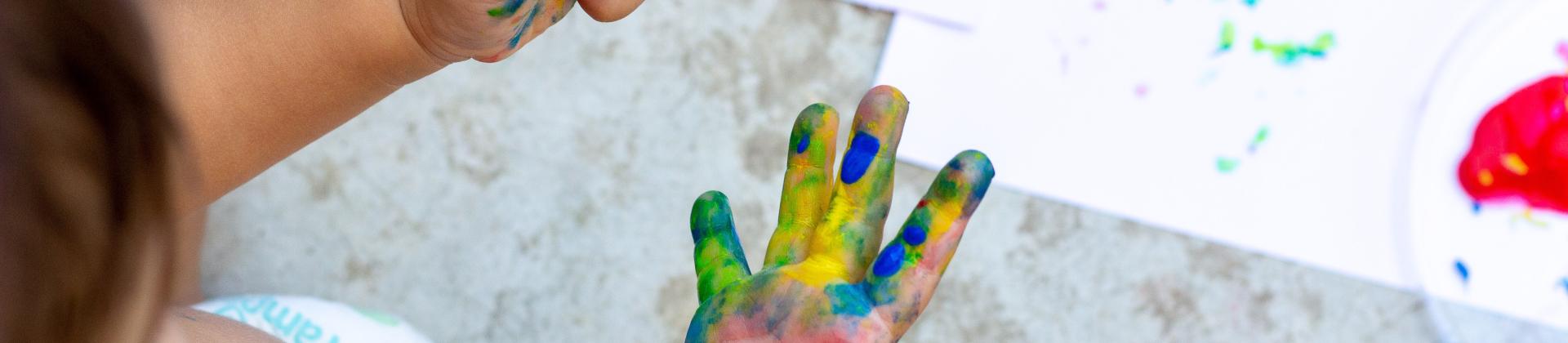 A small child is painting. You can see only their hands covered in pink, blue, yellow and green paint.