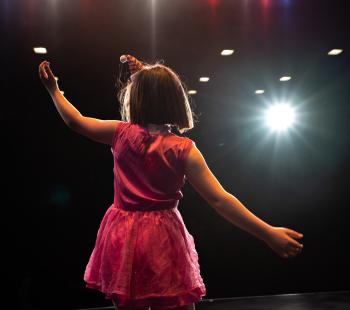 A girl in a pink dress and bobbed brown hair performs onstage with her arms outstretched.