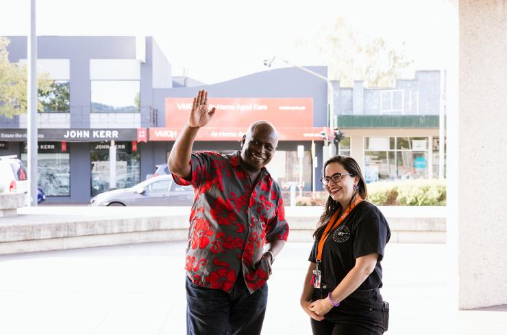 Man in red shirt waving outside Moe Library, standing next to a woman in a black shirt, smiling.