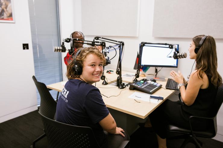 People sitting at microphones in a podcast studio. One person is smiling at the camera. Equipment is on the table in front of them.