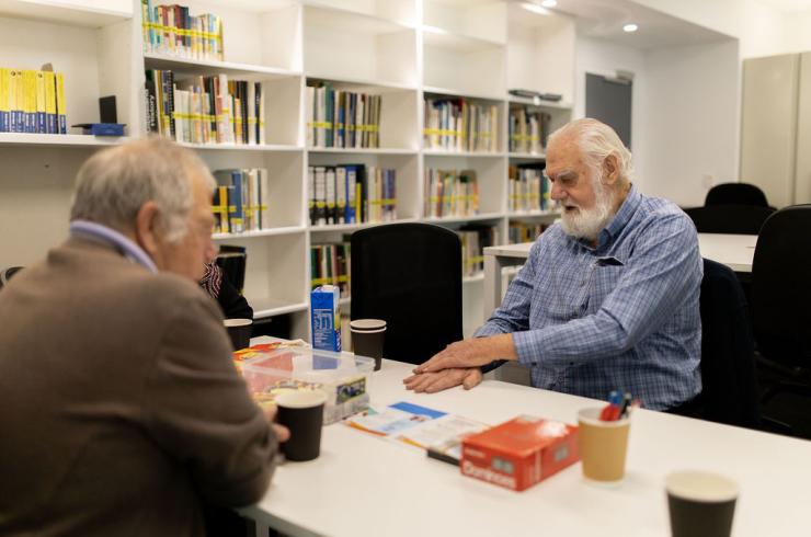 Two older white men sitting opposite each other at a table in the library, talking.