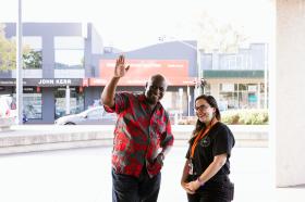Man in red shirt waving outside Moe Library, standing next to a woman in a black shirt, smiling.