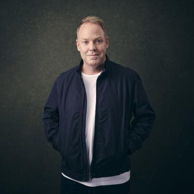 Peter Helliar looks directly at the camera. He has blonde hair and is wearing a white t-shirt, black jeans and a navy blue bomber jacket.