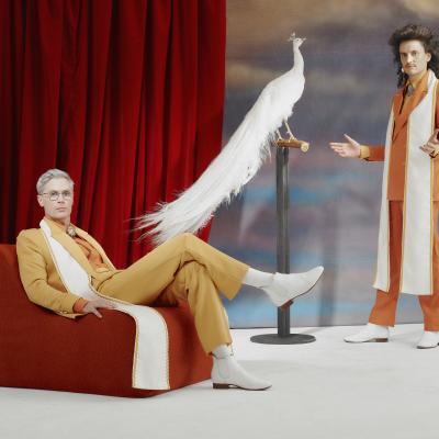 Two young men dressed in yellow and orange dapper suits pose with a white peacock