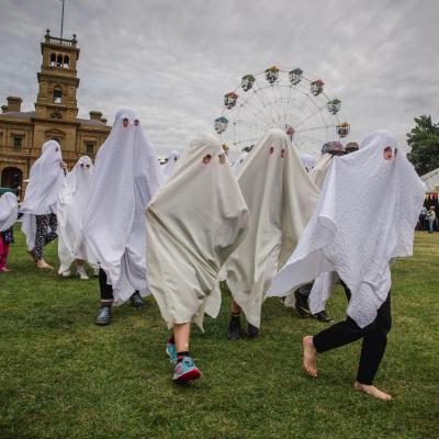 A group of silly young people run through a grassy park dressed up as ghosts. They have white sheets thrown over their heads with round eye holes cut out so they can see.