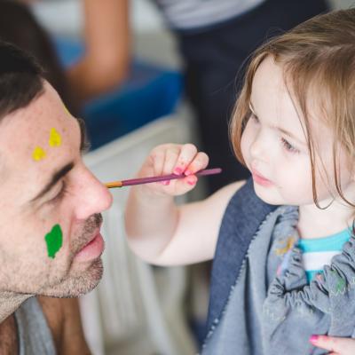 a small blonde girl paints her fathers face with a paint brush. He has yellow spots on his forehead and a green blob on his left cheek.