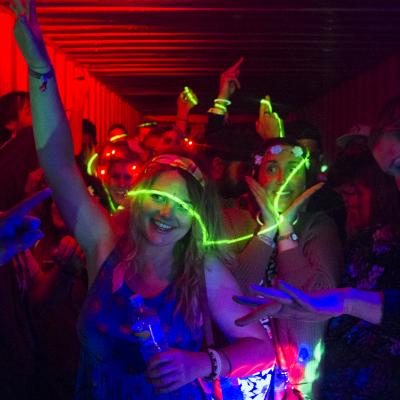 Young adult girls are going wild dancing with glow sticks and dim red lighting. They're at a ten minute dance party inside of a shipping container with a packed crowd.