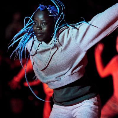 A girl with black skin hip hop dances with great energy. There are other young dancers in the background lit with vivid red lighting.