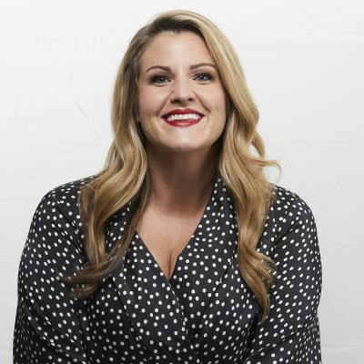 Nikki Britton is smiling at the camera. She has long blonde wavy hair and a black blouse with a fine white dotty pattern.