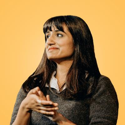 Vidya Rajan looks into the distance to the left. She has long, straight rich brown hair with a fringe. She is wearing a dark green jumper with a white collar poking out. She poses in front of a yellow background.