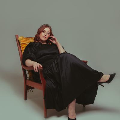 Meredith sits on a vintage chair in a black silk dress. She is classy.