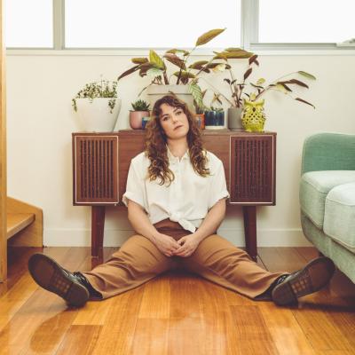 Darcy Fox sits on a wooden floor with taupe slacks and a cream shirt. She looks wistful.