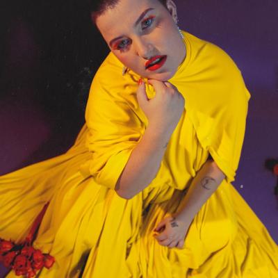 Emma sits against a purple background resting her chin on her hand. She wears a spectacular yellow jumpsuit with neon make up and has a shaved head.