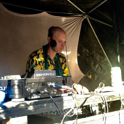 DJ Jaime Mack spins tracks on the decks. He is bald and fair skinned. He is wearing a tropical shirt.